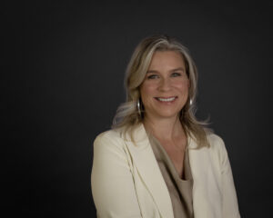 White woman, mid 40s, long blonde hair and silver hoop earrings wearing a beige blazer over a gold blouse.