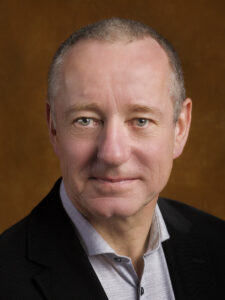 White man, 50s with short grey hair wearing a grey shirt and black blazer