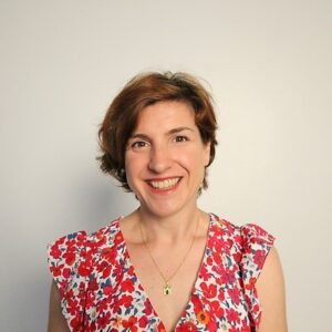 White woman, early 40s, short red/brown hair wearing a red floral blouse and gold necklace