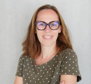 White woman, 40s, long light brown hair wearing glasses and earrings with a green polk-dot t-shirt